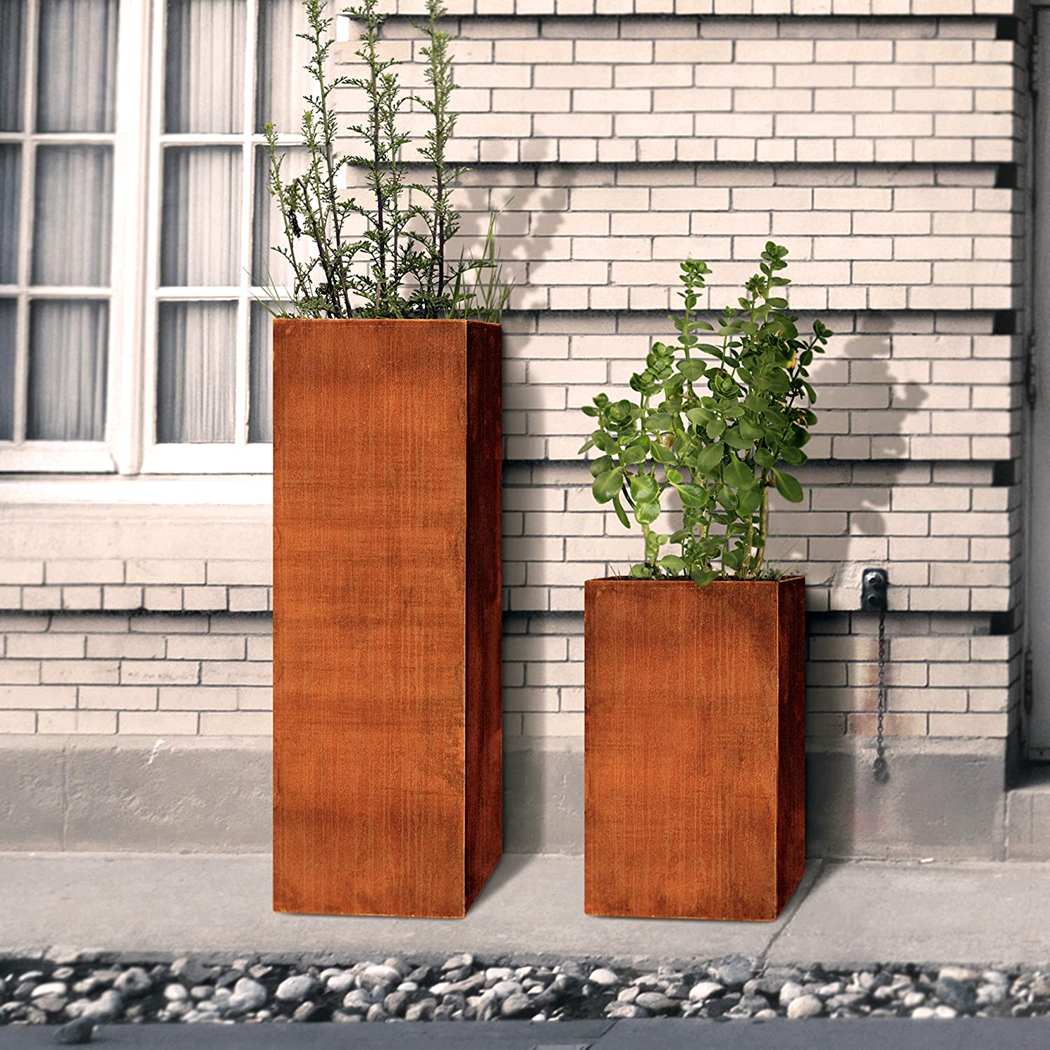 Two tall Corten Steel planters with foliage in them against a white brick wall.